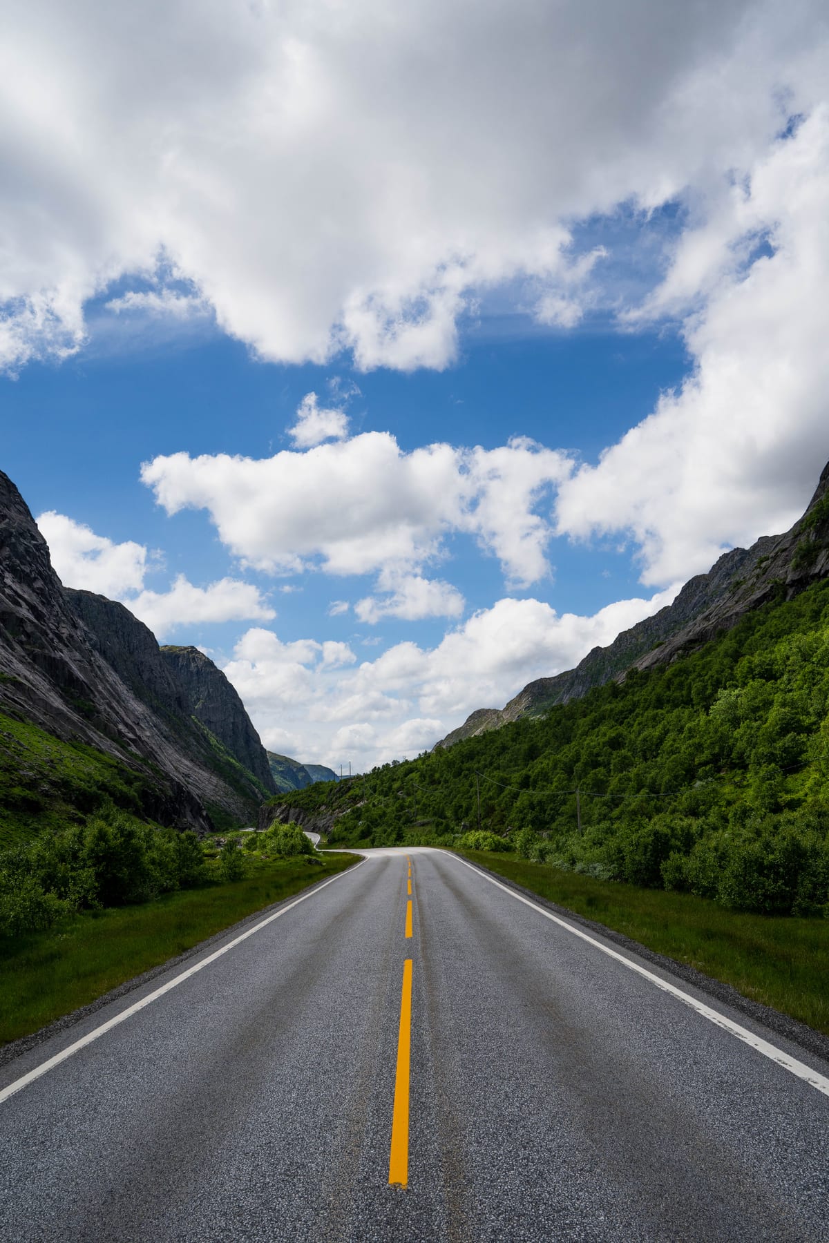 Eine gerade Straße mit gelber Mittellinie führt zwischen grünen Berghängen unter einem blauen Himmel mit weißen Wolken hindurch.