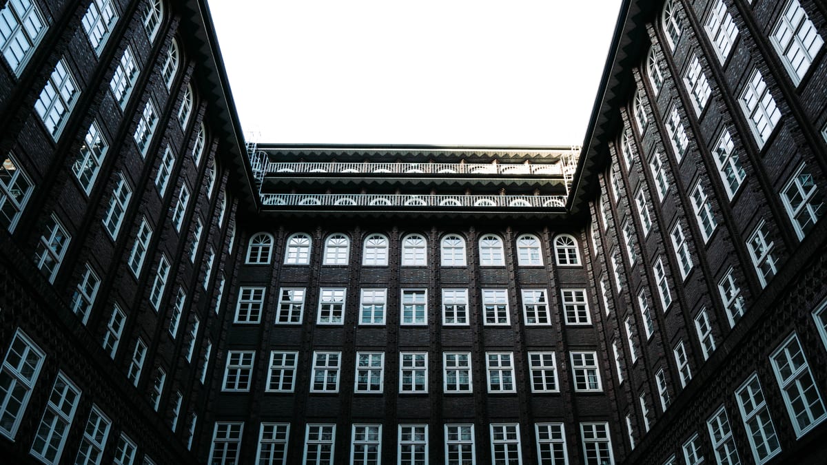 An upward view of dark brick facades and windows around a courtyard against a bright sky.