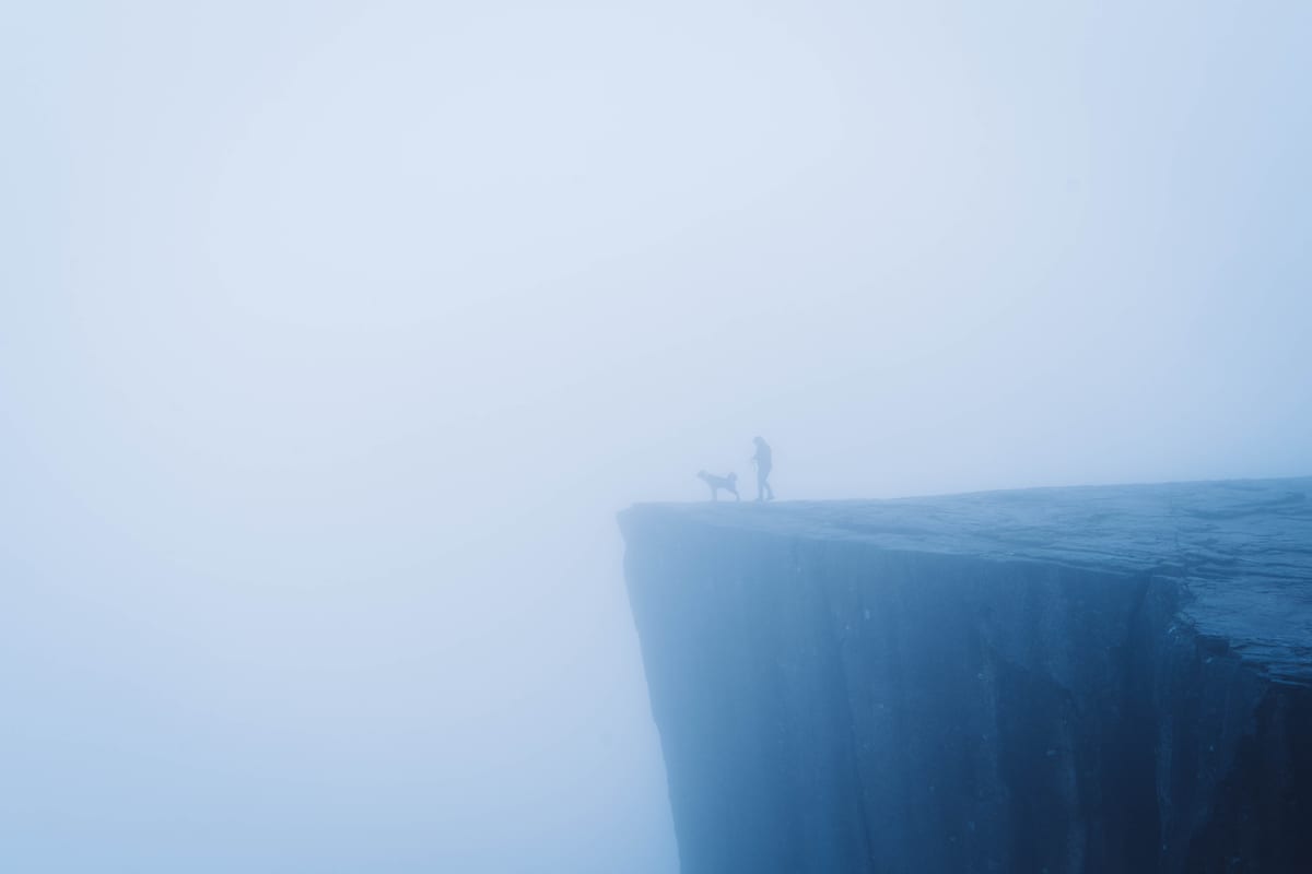 Kleine menschliche Silhouetten auf einer steilen Felsplattform, die im dichten Nebel verschwindet.