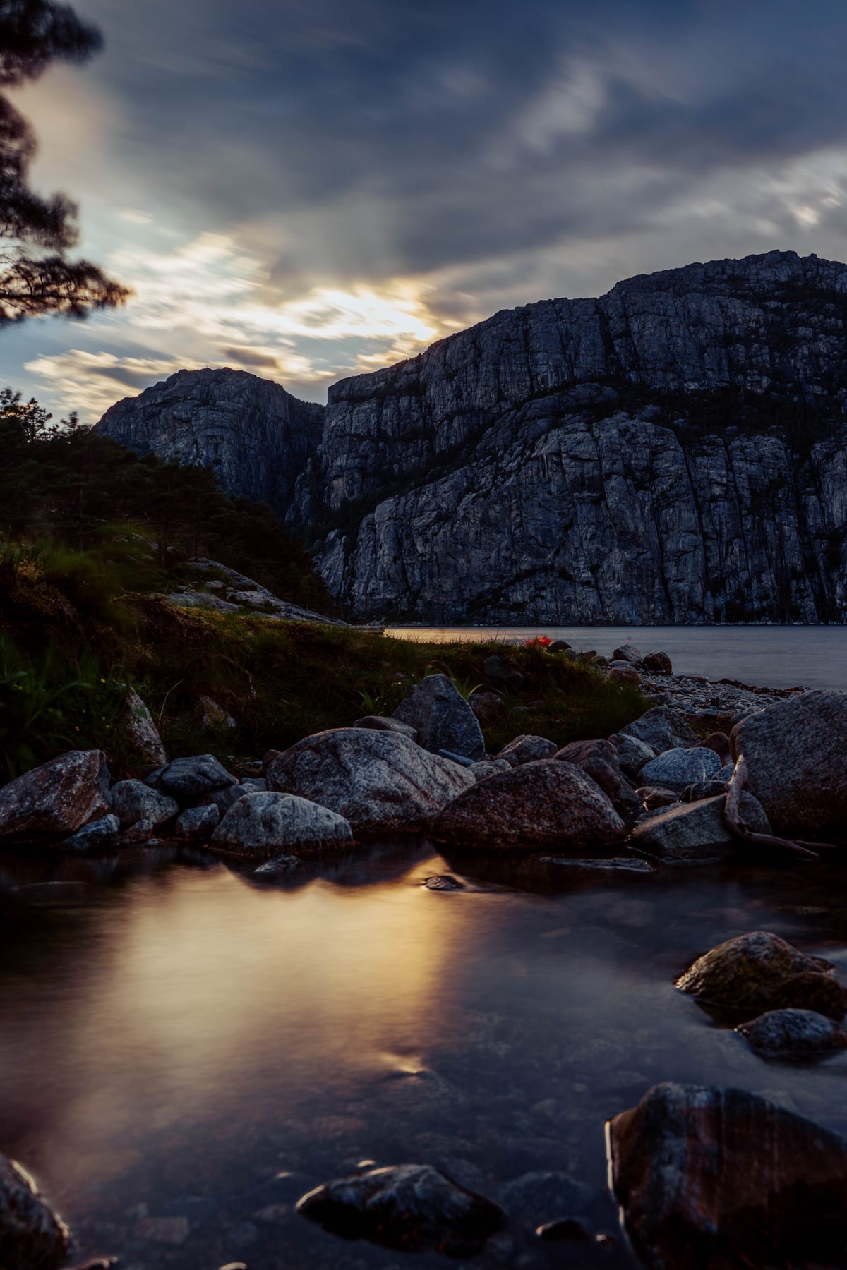 Wasser fließt zwischen glatten Felsen unter einer steilen Felswand und einem wolkigen Abendhimmel.