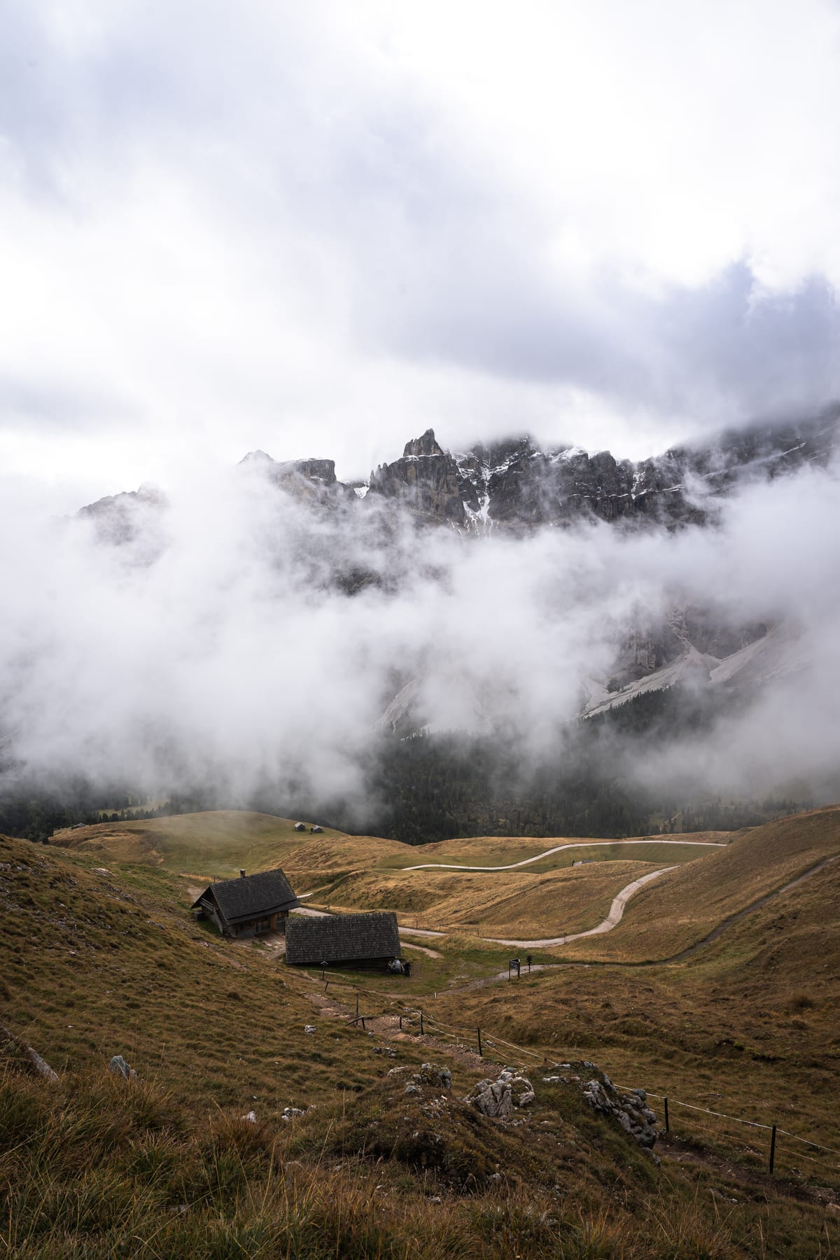 Two huts on an alpine meadow beneath cloud-covered rocky peaks.