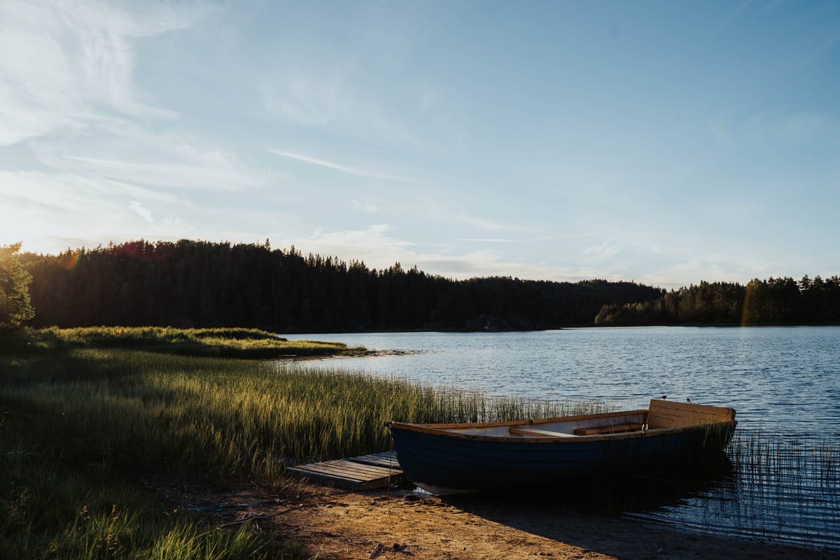 Ein Ruderboot liegt an einem Holzsteg am stillen See, umgeben von Schilf und bewaldeten Ufern im Abendlicht.