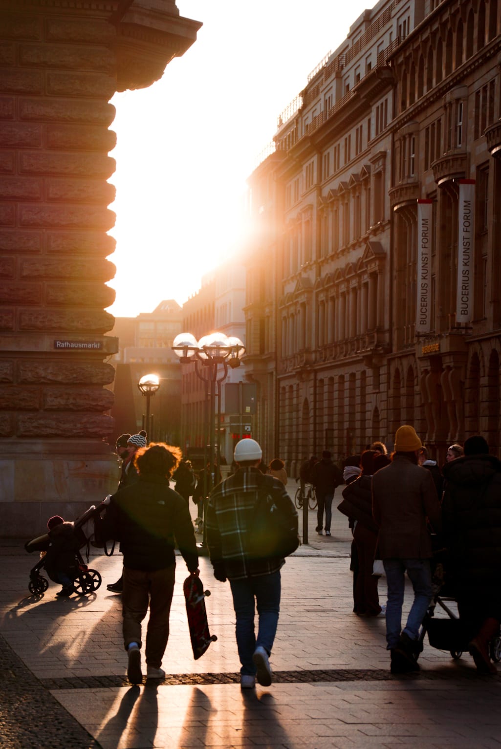 People walk along a street between tall buildings, backlit by the golden light of the low sun.