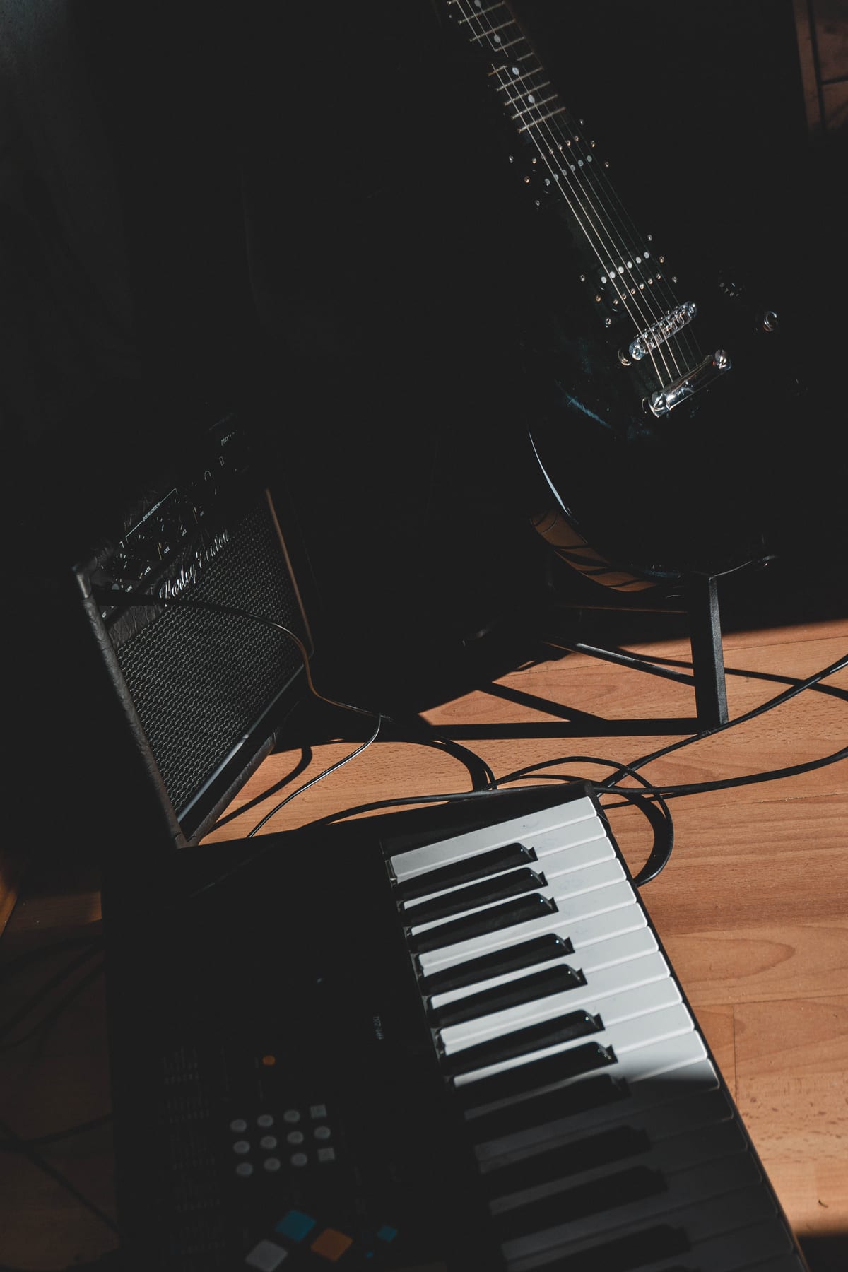 An electric guitar, an amplifier and a keyboard on a wooden floor in dim light.