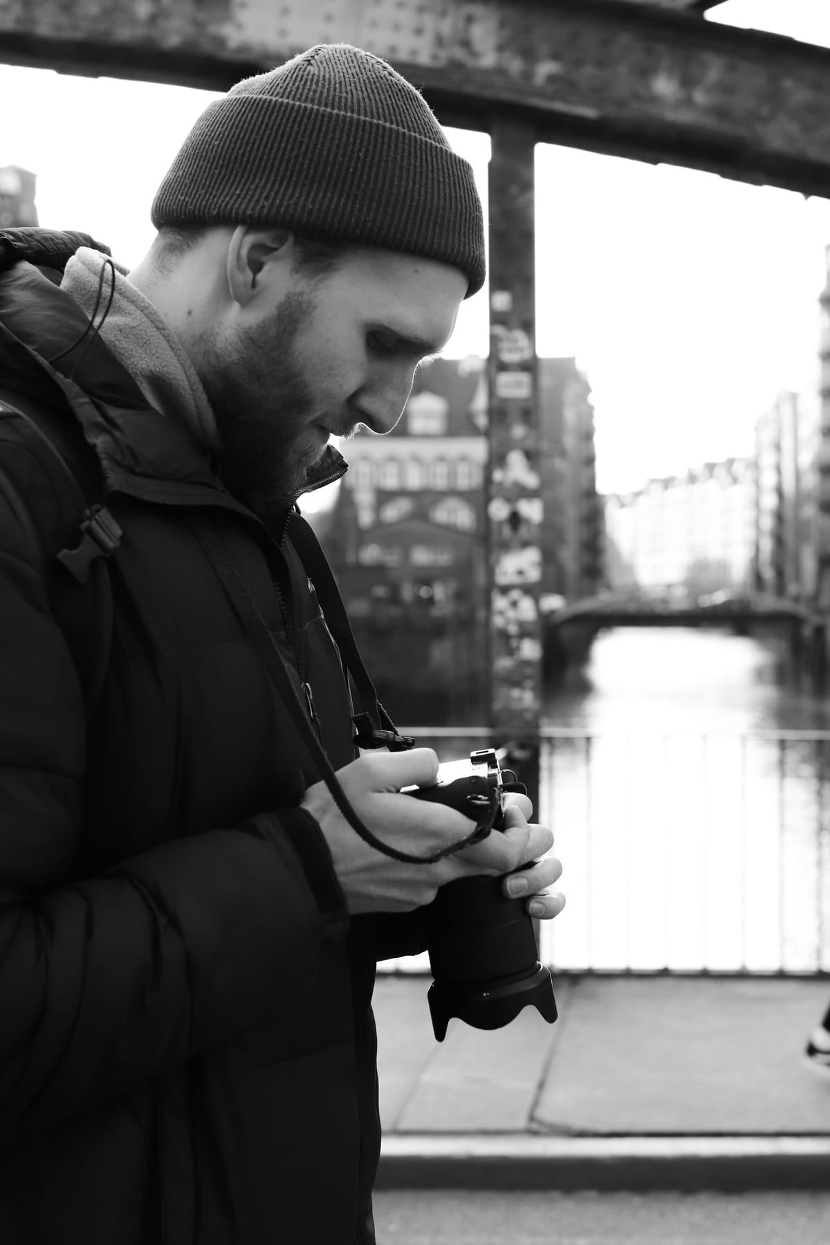 A black and white photograph of a man wearing a beanie, looking down at his camera beside the water.