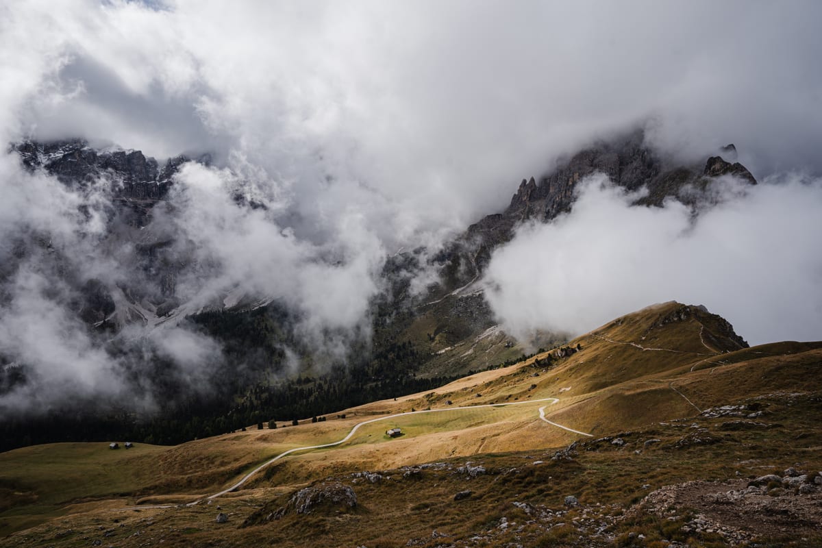 Sunlight on an alpine meadow with winding paths beneath dense clouds and dark mountainsides.