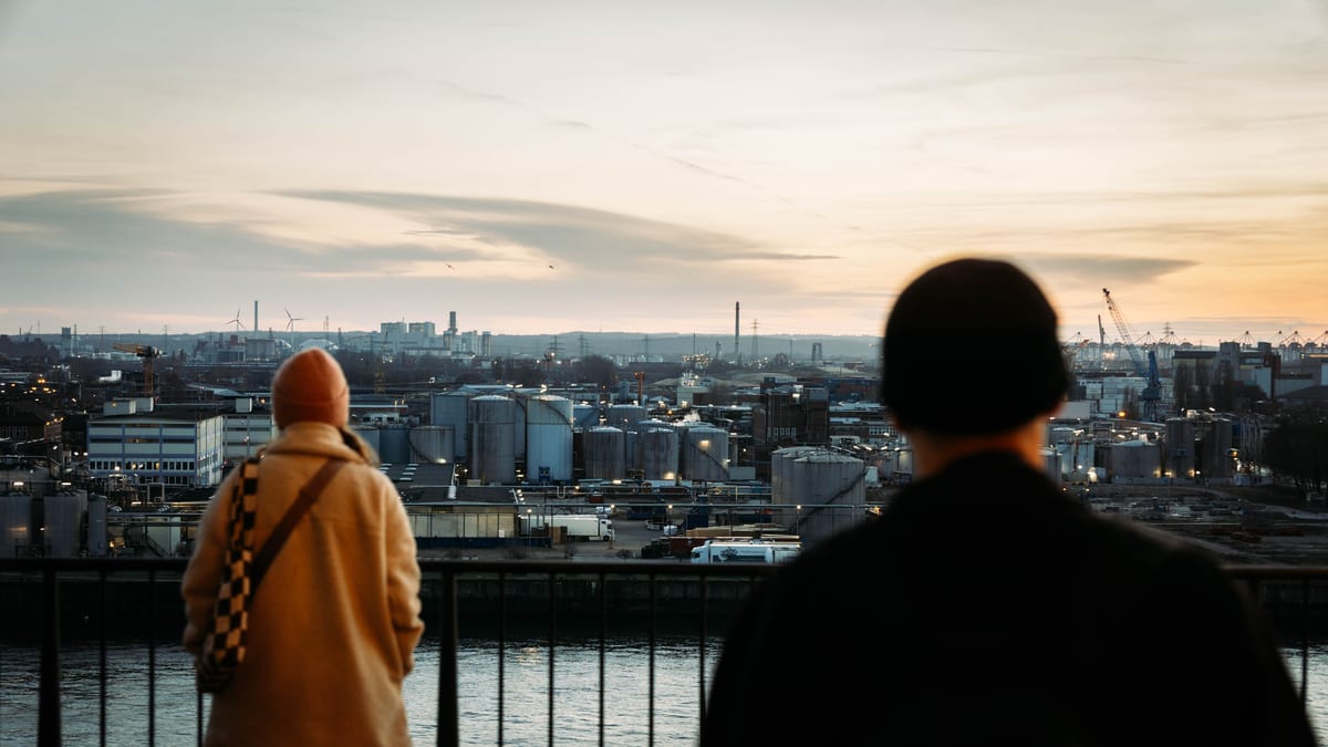 Two people at a railing overlooking the water and harbour in evening light.