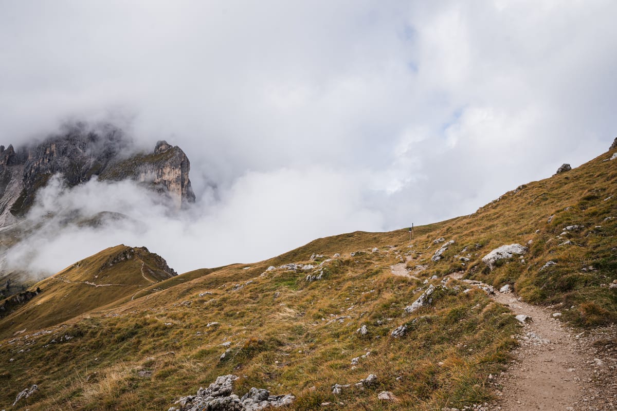 Ein Wanderweg führt über einen grasigen Hang zu Felsgipfeln im Nebel.