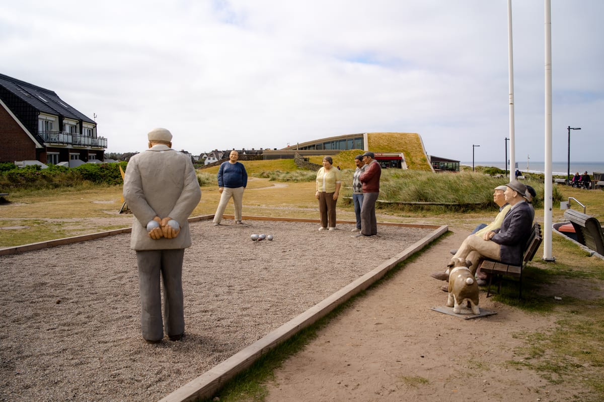 A group of sculptures beside a boules court between houses and grassy dunes.