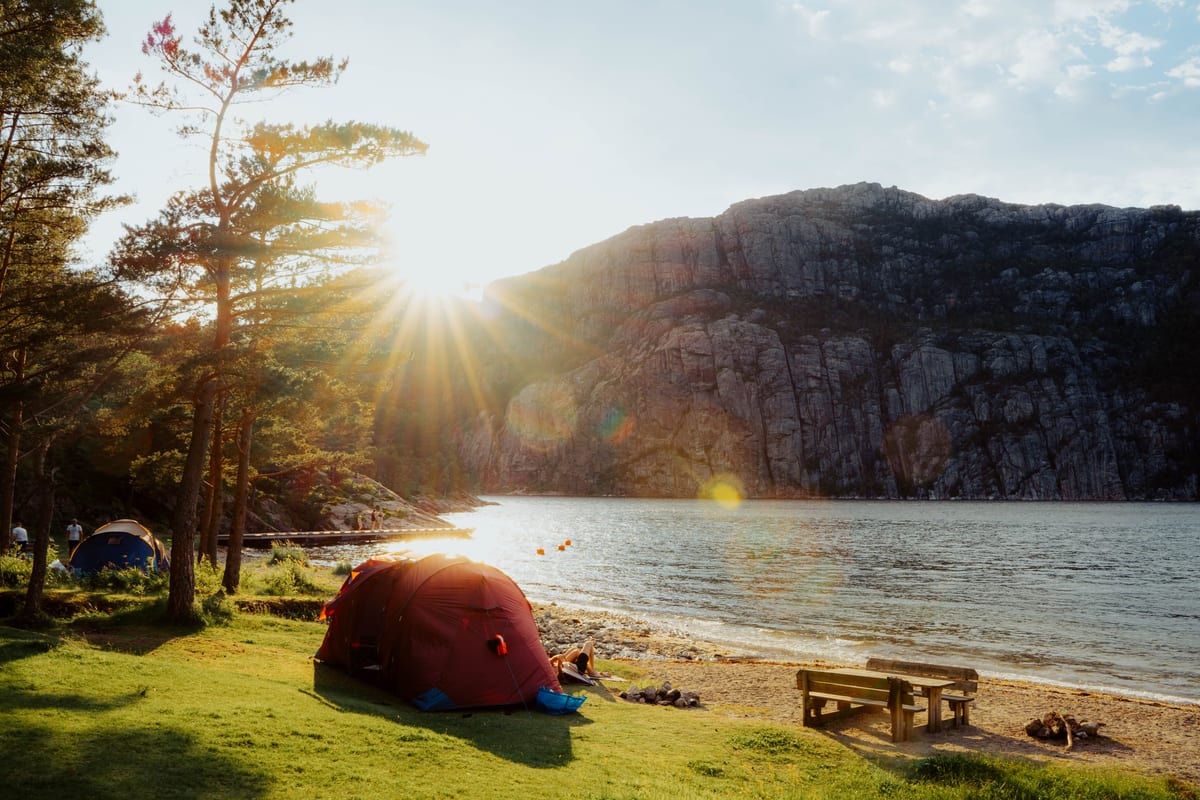 Ein rotes Zelt auf einer Wiese am Wasser, mit Kiefern, Picknickbänken und hohen Felswänden im Sonnenlicht.