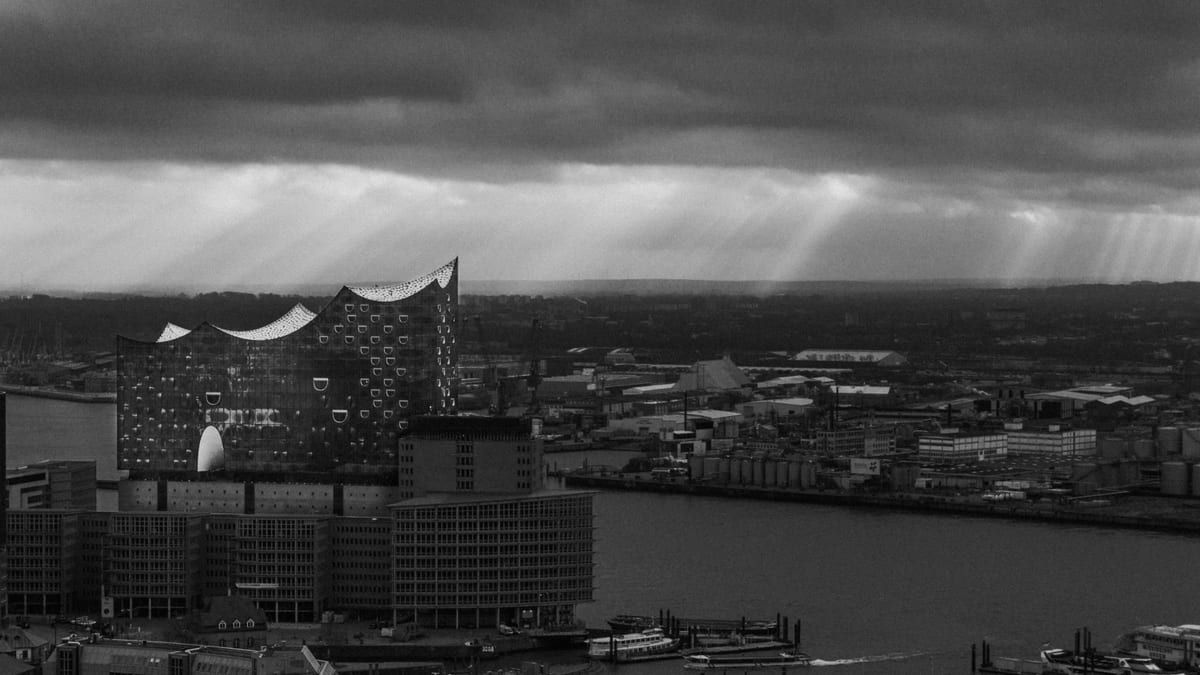 A black and white view of the Elbphilharmonie and Hamburg harbour beneath dark clouds with rays of sunlight.
