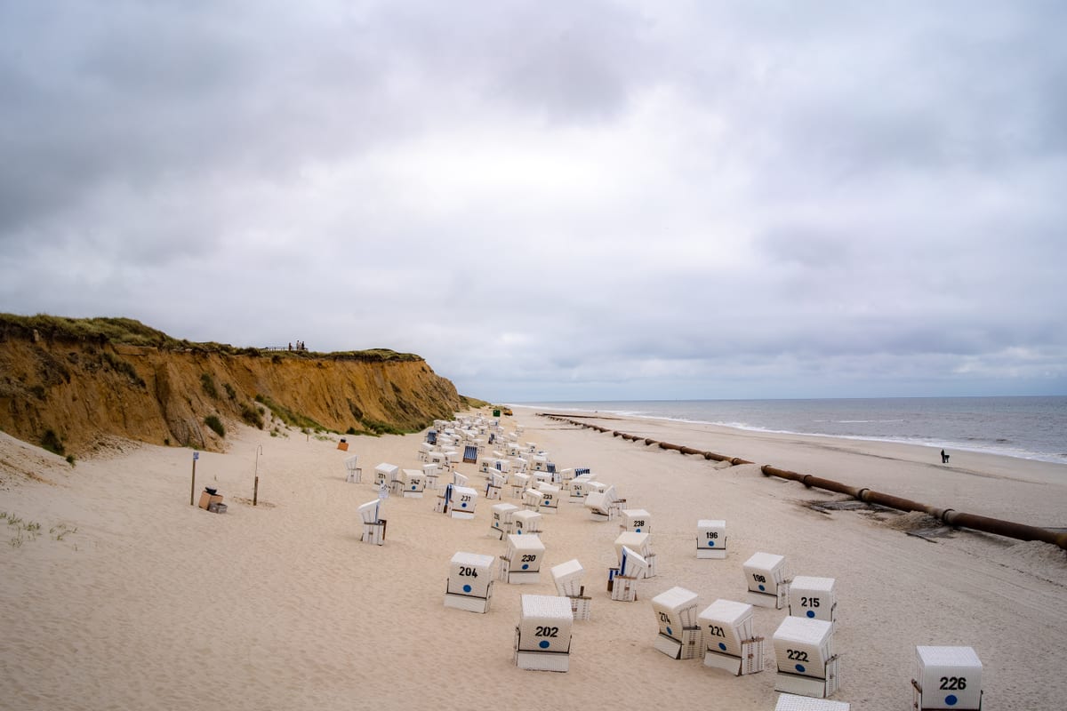 White hooded beach chairs on a sandy beach beside a steep coastal bluff under a cloudy sky.