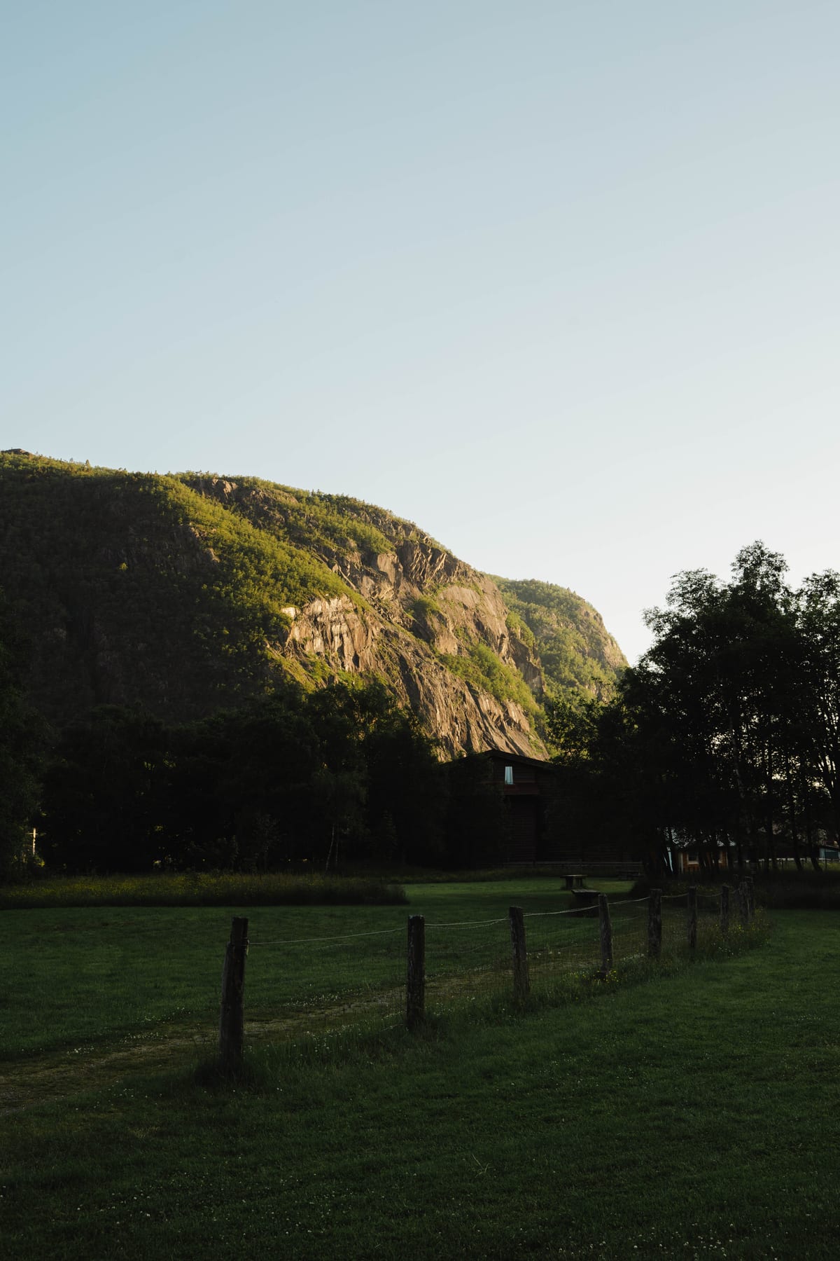 Abendsonne beleuchtet einen grünen Berghang über einer schattigen Wiese mit Holzzaun.